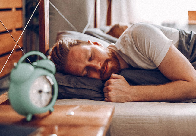 Man ligt te slapen in bed met een wekker naast hem op het nachtkastje